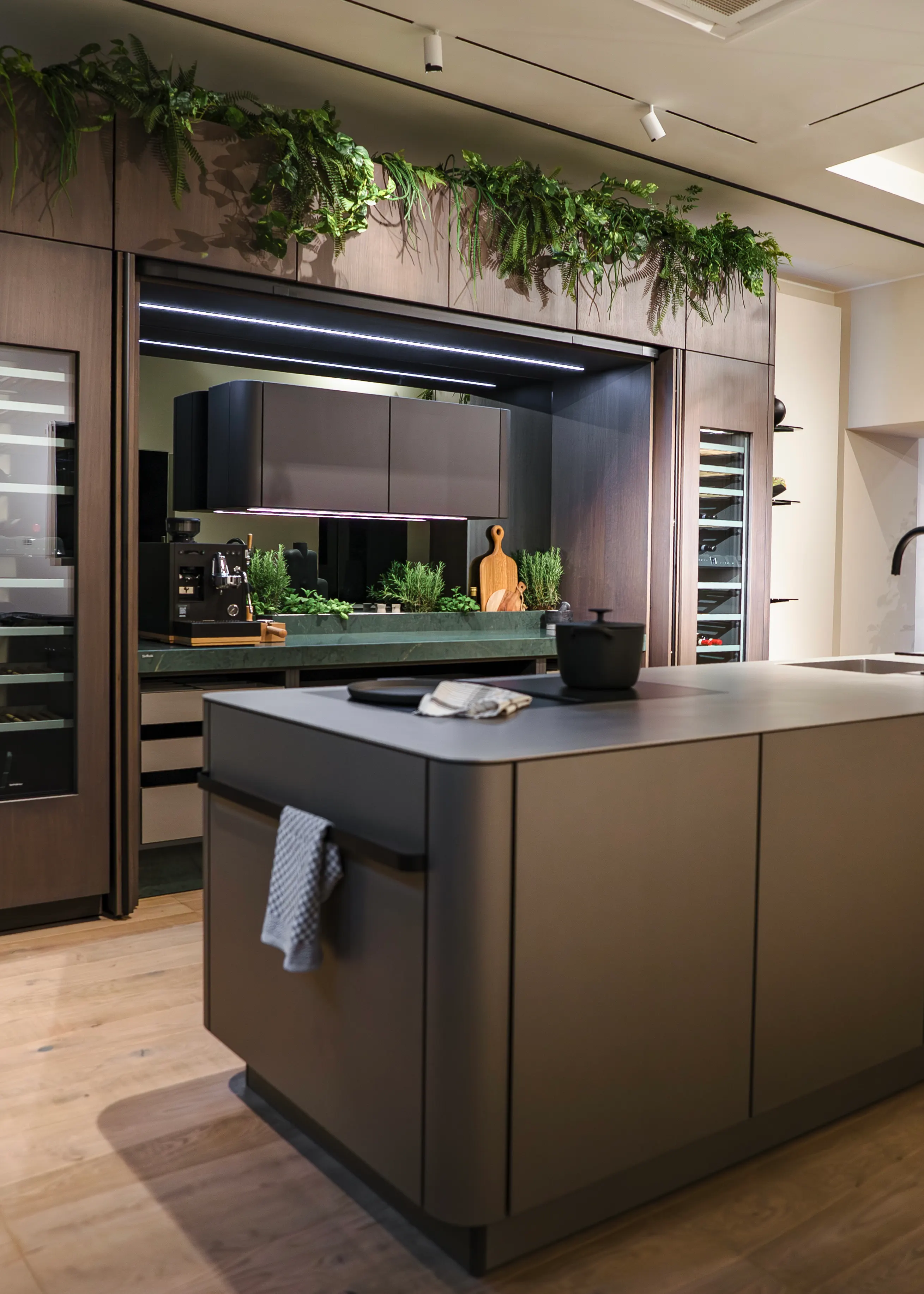 Moderne SieMatic SG6 Luxusküche mit einer dunkelbraunen, skulpturalen Kochinsel, eleganten Holzfronten, integriertem Weinkühlschrank und grünen Pflanzen als Dekoration auf den Oberschränken.