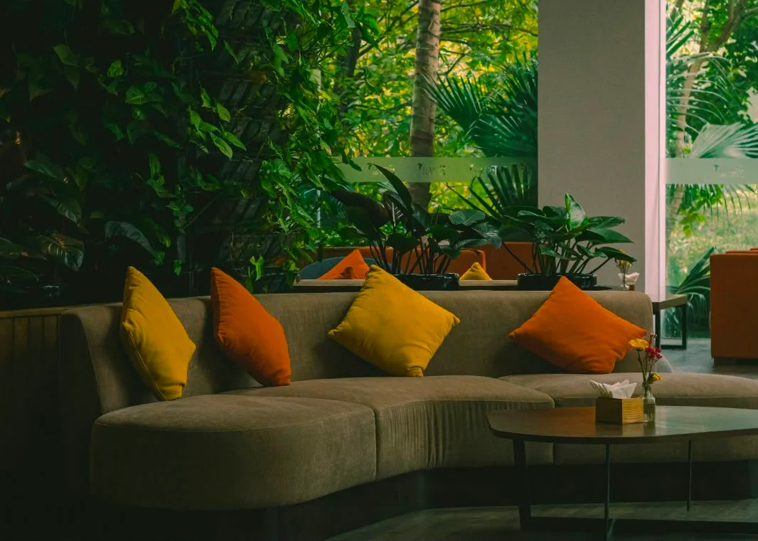 Luxuriöses geschwungenes Sofa in Erdtönen mit gelben und orangefarbenen Kissen in einem biophilen Wohnzimmer voller grüner Zimmerpflanzen.