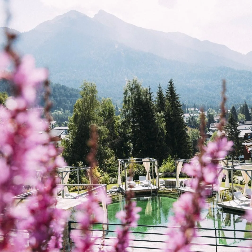 Naturbadeteich im Alpin Resort Sacher – Luxus-Wochenend-Escape Naturbadeteich im Außenbereich des Alpin Resort Sacher in Seefeld mit Liege-Pavillons, umrahmt von lila Blumen.