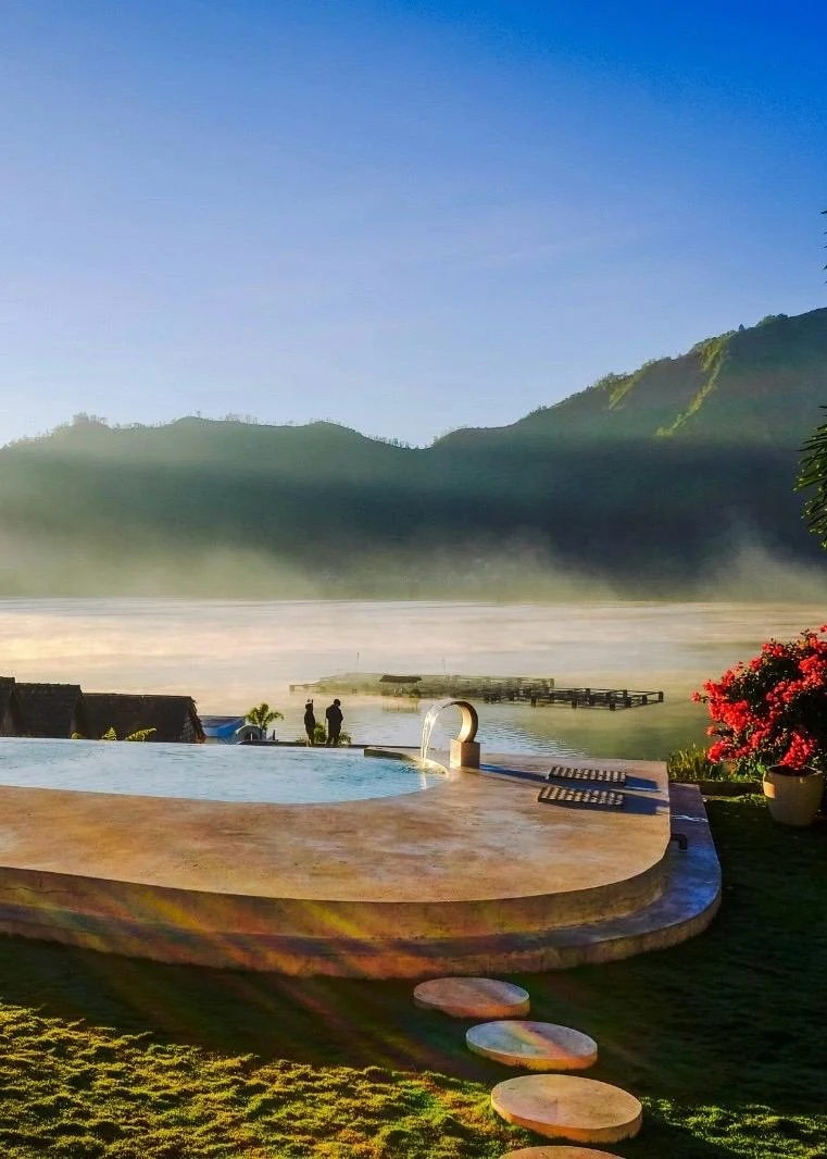 Ein organisch geformter Infinity-Pool aus Naturstein mit Blick auf einen nebligen See und Berge im warmen Morgenlicht des Sonnenaufgangs.