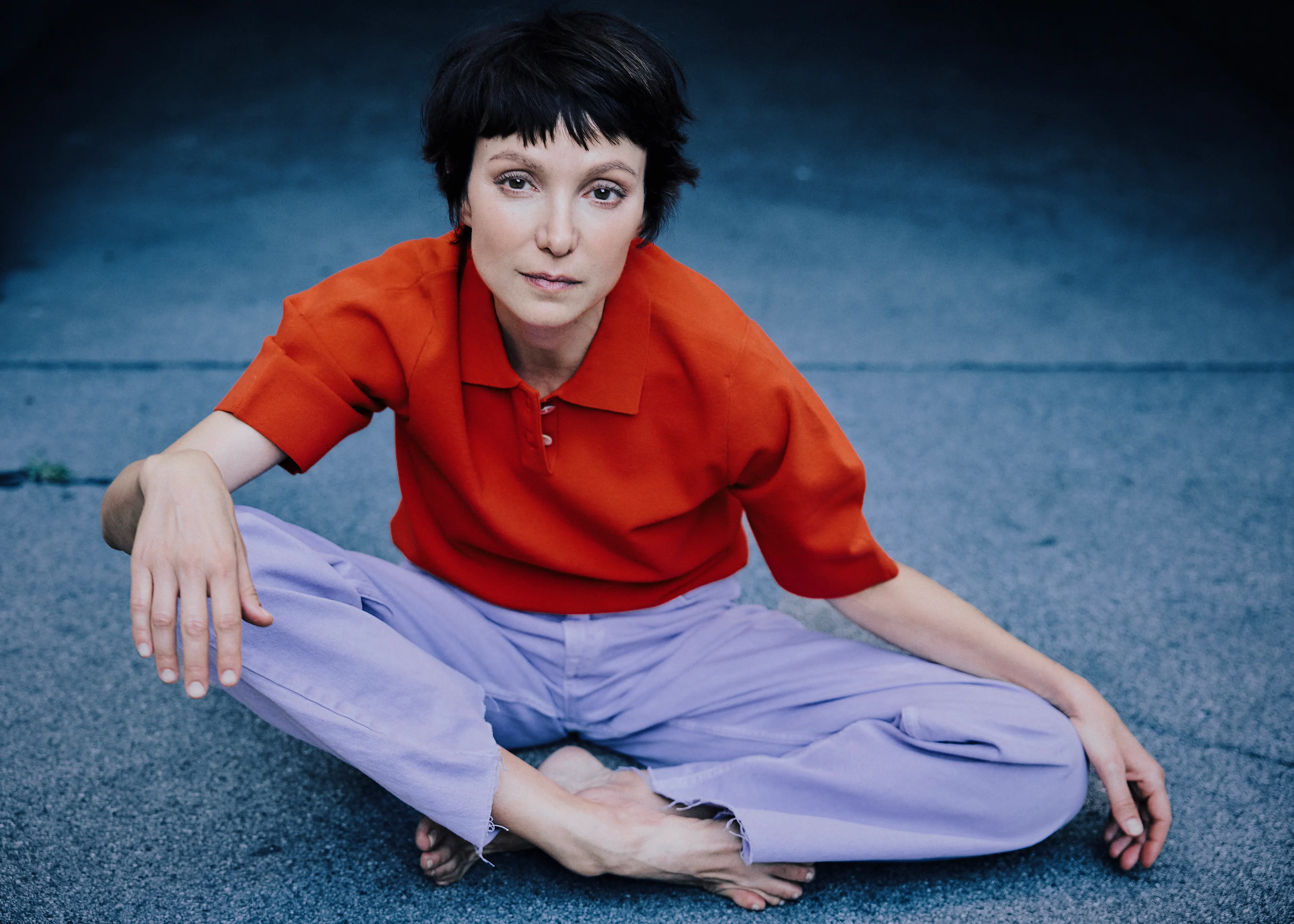 Schauspielerin Julia Koschitz sitzt in rotem Poloshirt und fliederfarbener Hose auf dem Boden.