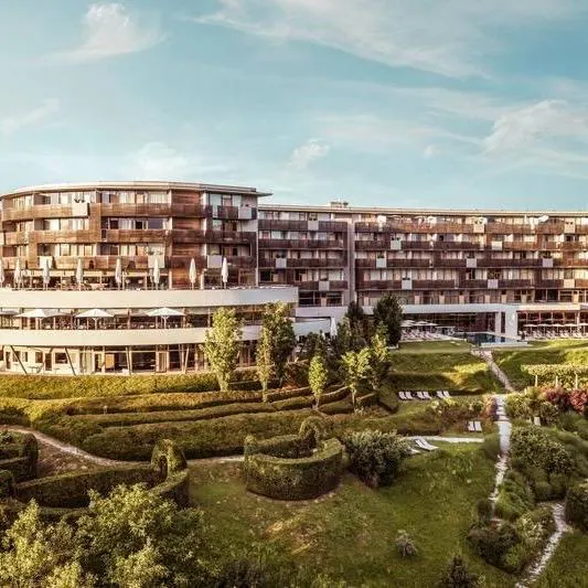 Außenansicht des modernen Falkensteiner Balance Resort Stegersbach mit seiner geschwungenen Holz- und Glasfassade, umgeben von einer gepflegten, grünen Gartenanlage mit Heckenstrukturen und Liegewiesen unter blauem Himmel.