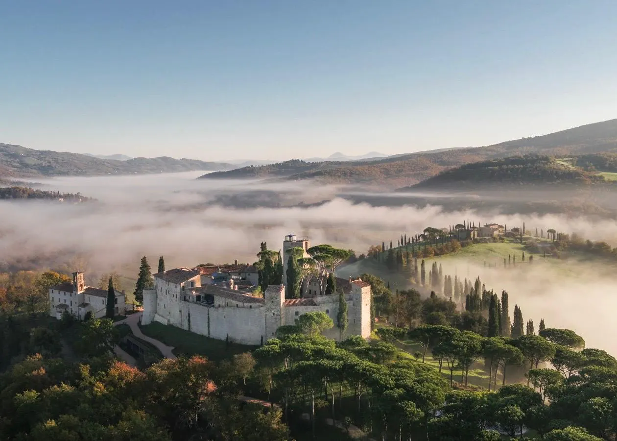 Vogelperspektive auf das historische Castello di Reschio in Umbrien, eingebettet in eine neblige Hügellandschaft mit Zypressen und Pinien.