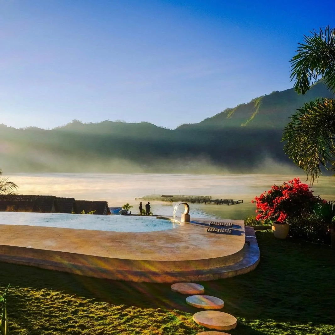 Ein organisch geformter Infinity-Pool aus Naturstein mit Blick auf einen nebligen See und Berge im warmen Morgenlicht des Sonnenaufgangs.