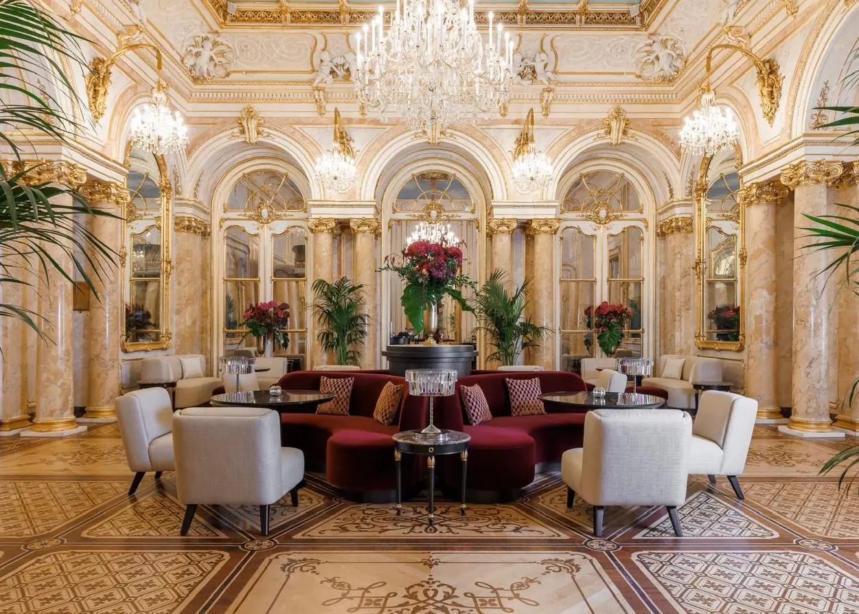 Prunkvoller historischer Saal im Luxushotel Palais Coburg in Wien mit weißen Sesseln, dunkelroten Sofas, Kristallleuchtern und Marmorsäulen.