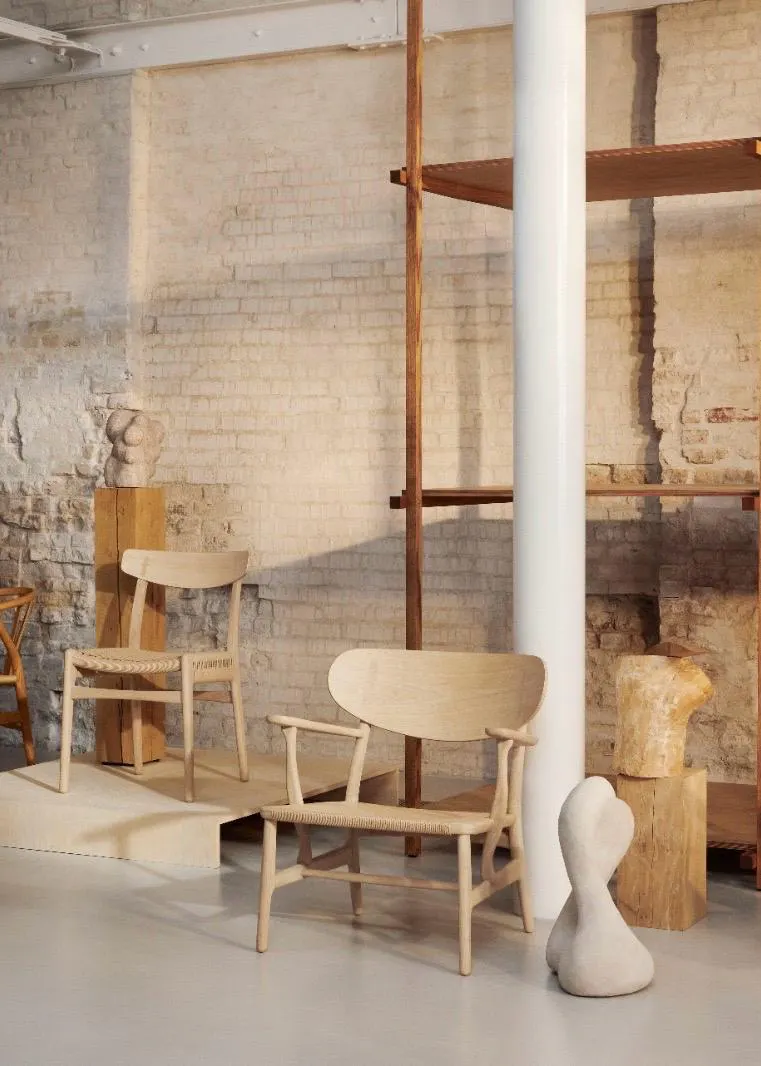 Ausstellungsraum im Loft-Stil mit minimalistischen Holzstühlen von Carl Hansen und Søn, darunter der CH24 Wishbone Chair, der CH22 Lounge Chair und der CH25 Sessel vor einer rustikalen Backsteinwand.