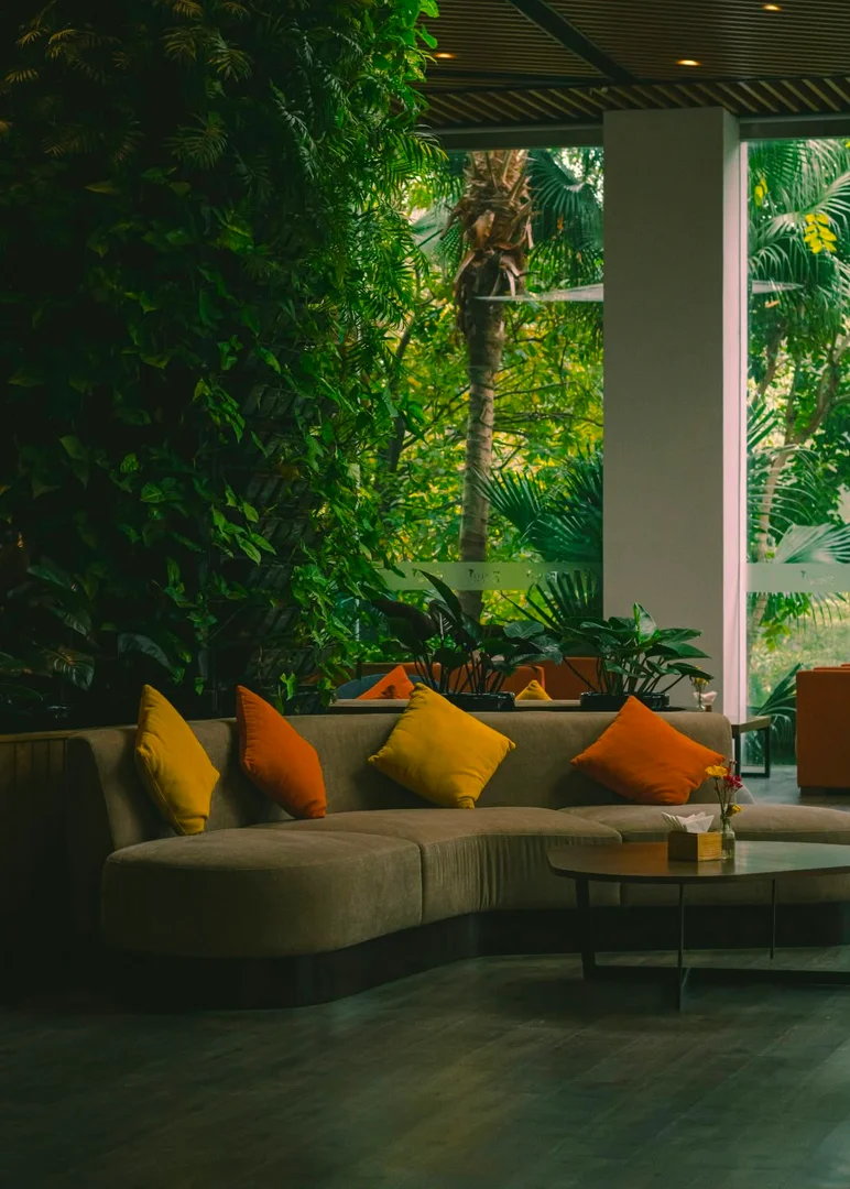 Biophilic Luxury: Fließende Übergänge zwischen Wohnraum und Natur Luxuriöses geschwungenes Sofa in Erdtönen mit gelben und orangefarbenen Kissen in einem biophilen Wohnzimmer voller grüner Zimmerpflanzen.