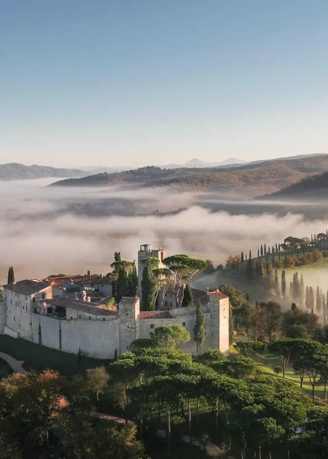 Vogelperspektive auf das historische Castello di Reschio in Umbrien, eingebettet in eine neblige Hügellandschaft mit Zypressen und Pinien.