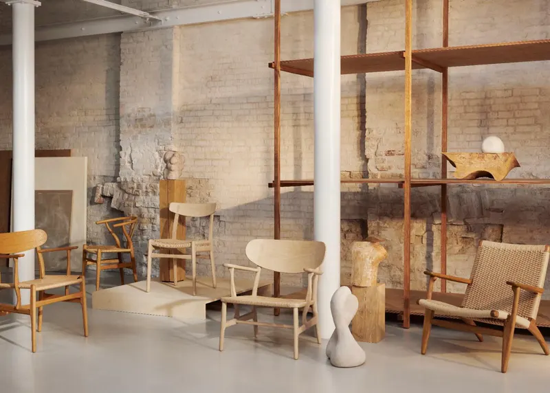 Ausstellungsraum im Loft-Stil mit minimalistischen Holzstühlen von Carl Hansen und Søn, darunter der CH24 Wishbone Chair, der CH22 Lounge Chair und der CH25 Sessel vor einer rustikalen Backsteinwand.