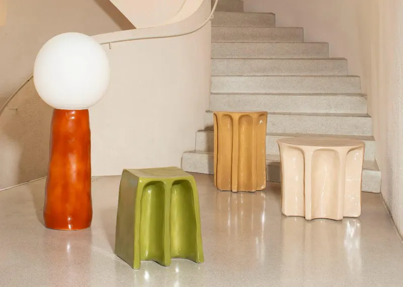 Ein minimalistischer Raum mit einer Treppe, in dem die Kokeshi Stehleuchte mit orangefarbenem Keramikfuß und die skulpturalen Chouchou Hocker und Beistelltische aus glasierter Keramik von Pulpo Products arrangiert sind.