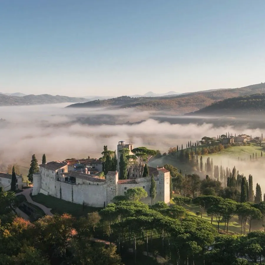 Castello di Reschio: Historische Architektur inmitten unberührter Natur Vogelperspektive auf das historische Castello di Reschio in Umbrien, eingebettet in eine neblige Hügellandschaft mit Zypressen und Pinien.