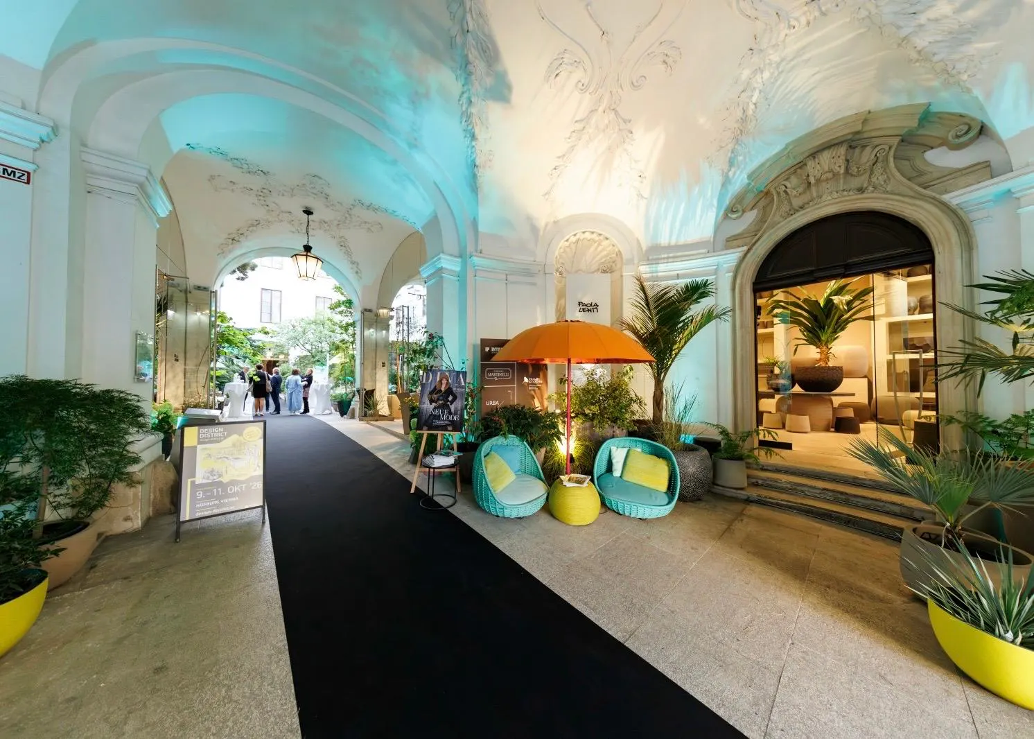 Exklusives Event-Setup im historischen Palais Ferstel in Wien mit schwarzem Teppich, türkisfarbenen Lounge-Sesseln unter einem orangen Sonnenschirm und einem Aufsteller des Design District.