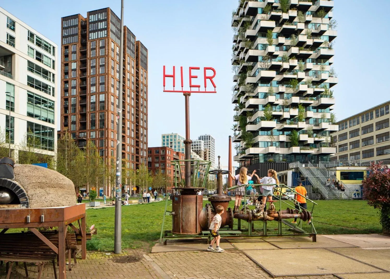 Kinder spielen auf einem grünen Platz im Kreativviertel Strijp-S in Eindhoven, im Hintergrund der bepflanzte Trudo Toren Wohnturm und die rote Leuchtschrift HIER.