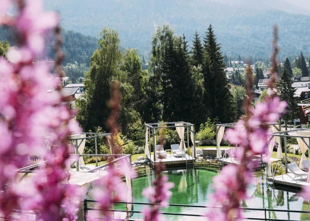Naturbadeteich im Alpin Resort Sacher – Luxus-Wochenend-Escape Naturbadeteich im Außenbereich des Alpin Resort Sacher in Seefeld mit Liege-Pavillons, umrahmt von lila Blumen.