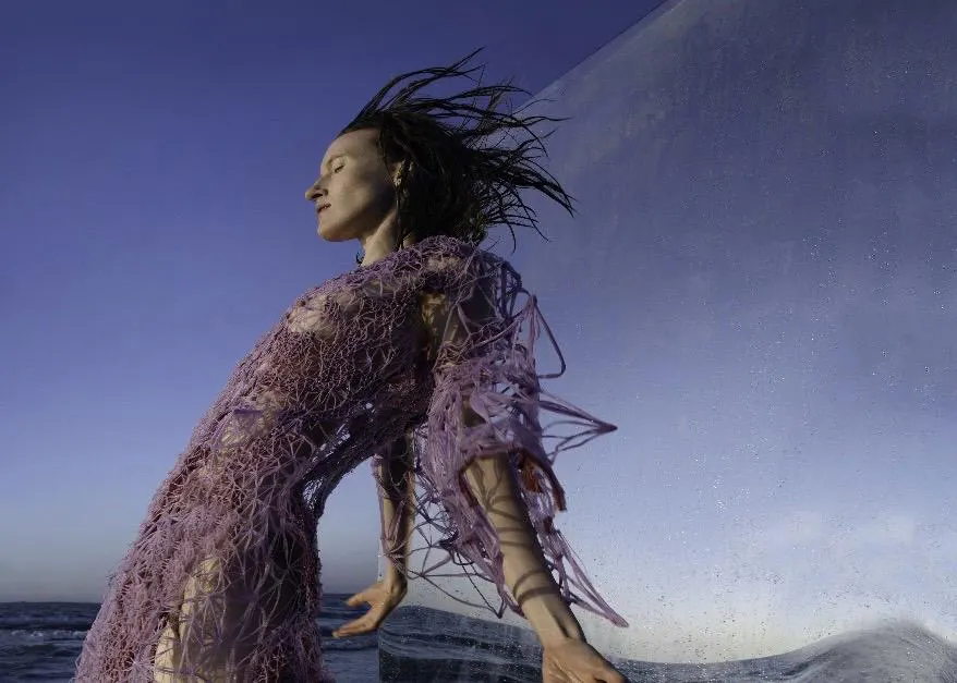 Model am Meer in einem lilafarbenen Kleid von Designerin Flora Miranda, das die feine Netzstruktur von 3D-Druck imitiert.