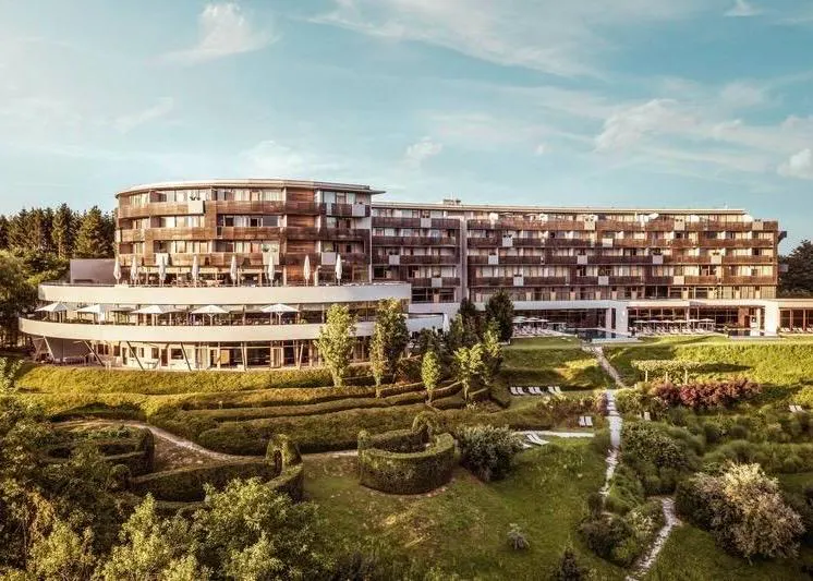 Außenansicht des modernen Falkensteiner Balance Resort Stegersbach mit seiner geschwungenen Holz- und Glasfassade, umgeben von einer gepflegten, grünen Gartenanlage mit Heckenstrukturen und Liegewiesen unter blauem Himmel.