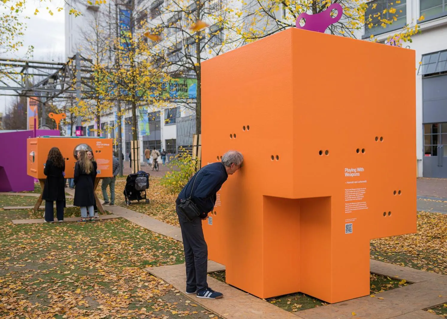 Ein Mann blickt durch Gucklöcher in eine große, orangefarbene Design-Installation in Würfelform mit der Aufschrift „Playing With Weapons“ auf der Dutch Design Week in Eindhoven.