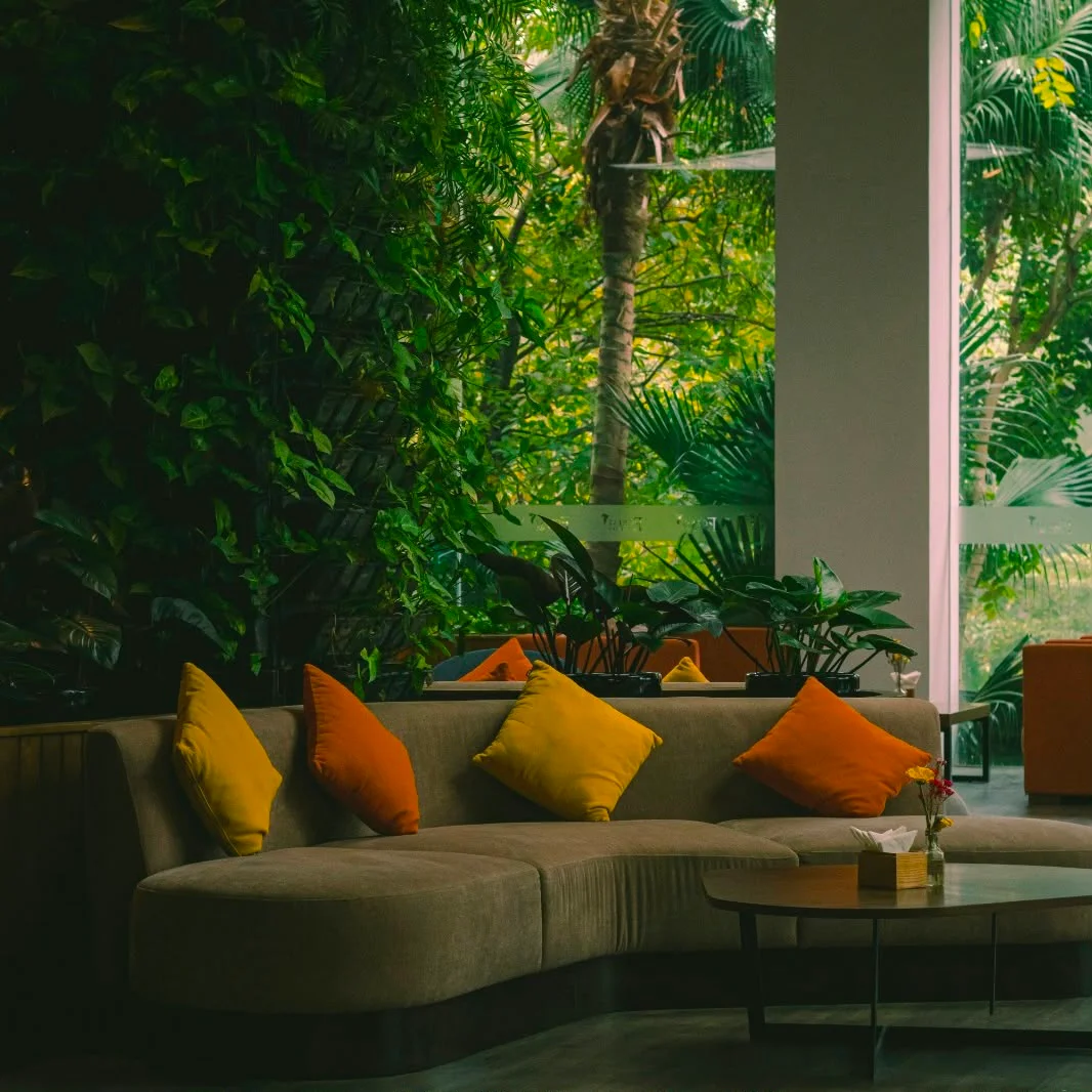 Biophilic Luxury: Fließende Übergänge zwischen Wohnraum und Natur Luxuriöses geschwungenes Sofa in Erdtönen mit gelben und orangefarbenen Kissen in einem biophilen Wohnzimmer voller grüner Zimmerpflanzen.