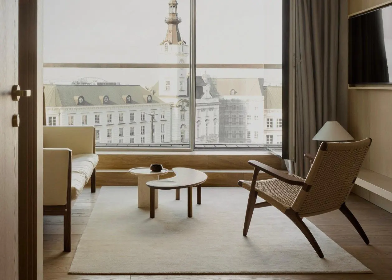 Minimalistisches Hotelzimmer im PURO Hotel Warschau Stare Miasto mit einem Hans Wegner CH25 Sessel, hellem Design-Sofa und großem Fenster mit Blick auf historische Gebäude in Warschau.