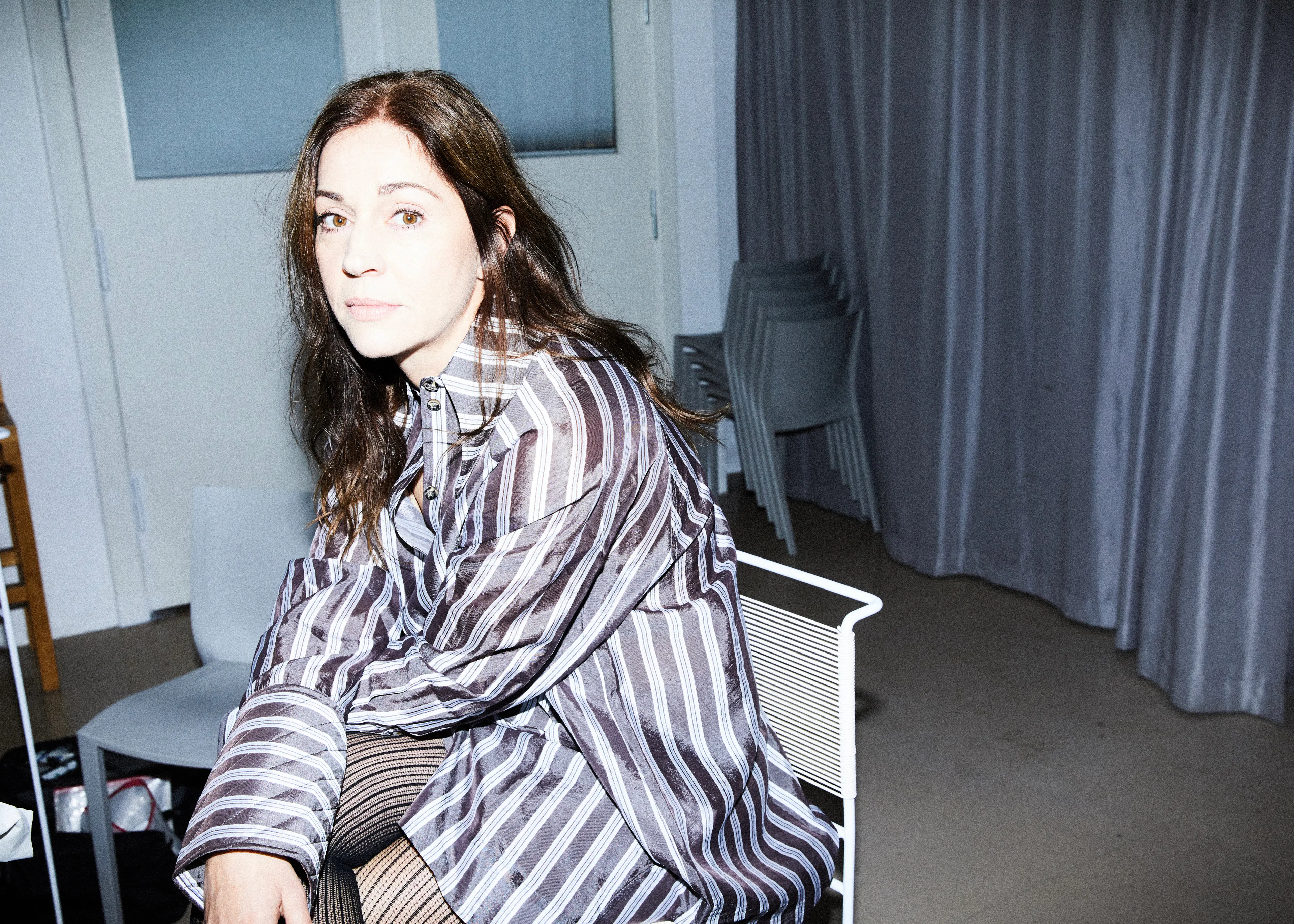 Porträt der Schauspielerin Ulrike C. Tscharre in gestreifter Bluse, sitzend auf einem weißen Designerstuhl im reduzierten Wohnbereich.