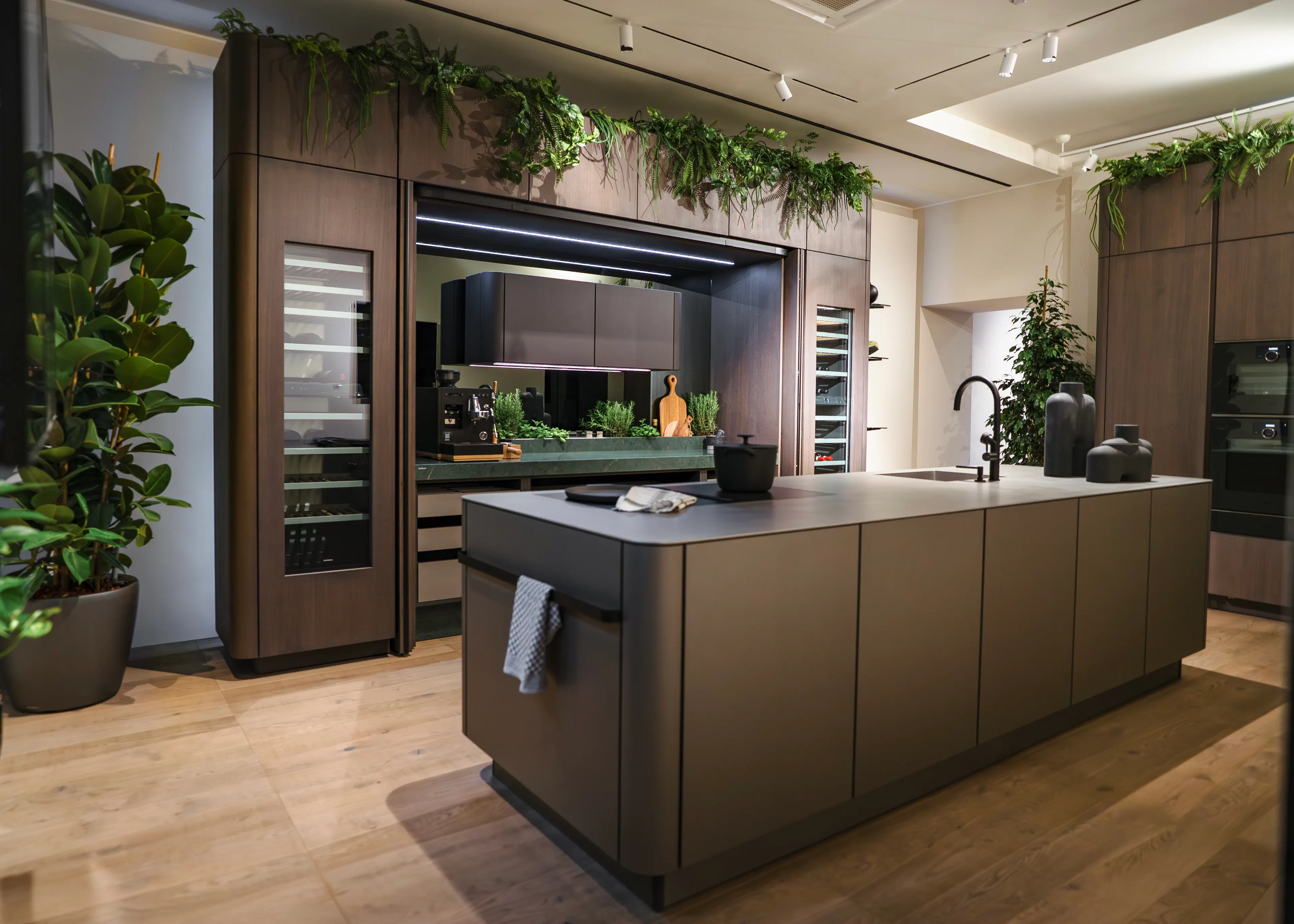 Moderne SieMatic SG6 Luxusküche mit einer dunkelbraunen, skulpturalen Kochinsel, eleganten Holzfronten, integriertem Weinkühlschrank und grünen Pflanzen als Dekoration auf den Oberschränken.