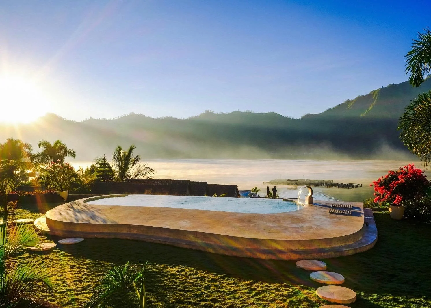 Ein organisch geformter Infinity-Pool aus Naturstein mit Blick auf einen nebligen See und Berge im warmen Morgenlicht des Sonnenaufgangs.